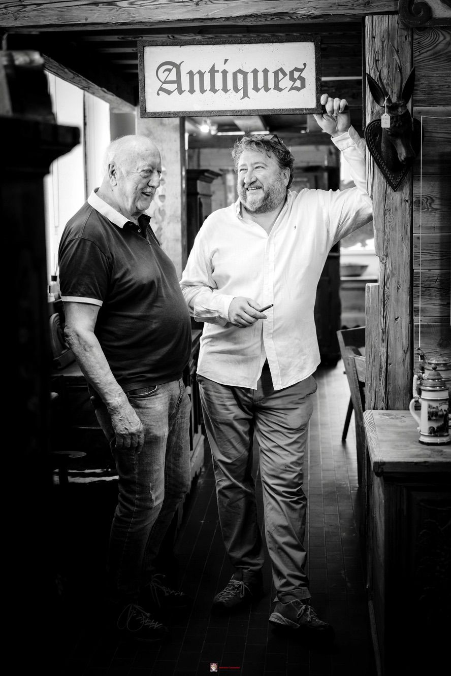 Leopoldo e Luca sorridono in un corridoio dell'esposizione, sotto un'insegna con la scritta Antiques