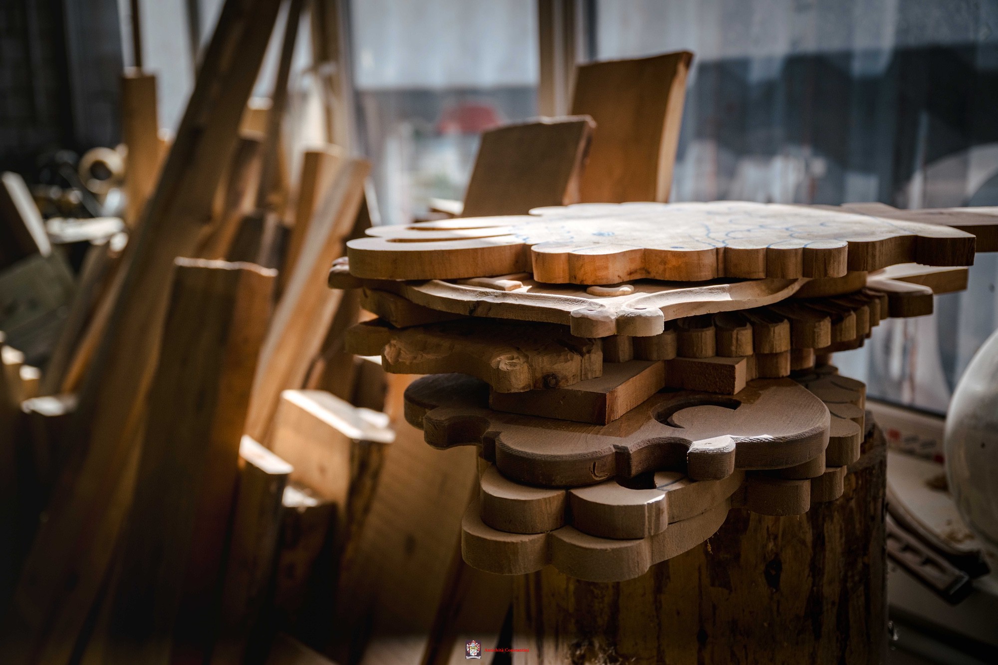 Pezzi di legno ritagliati a volute e impilati su un ceppo, accanto a tavole appoggiate alla finestra del laboratorio