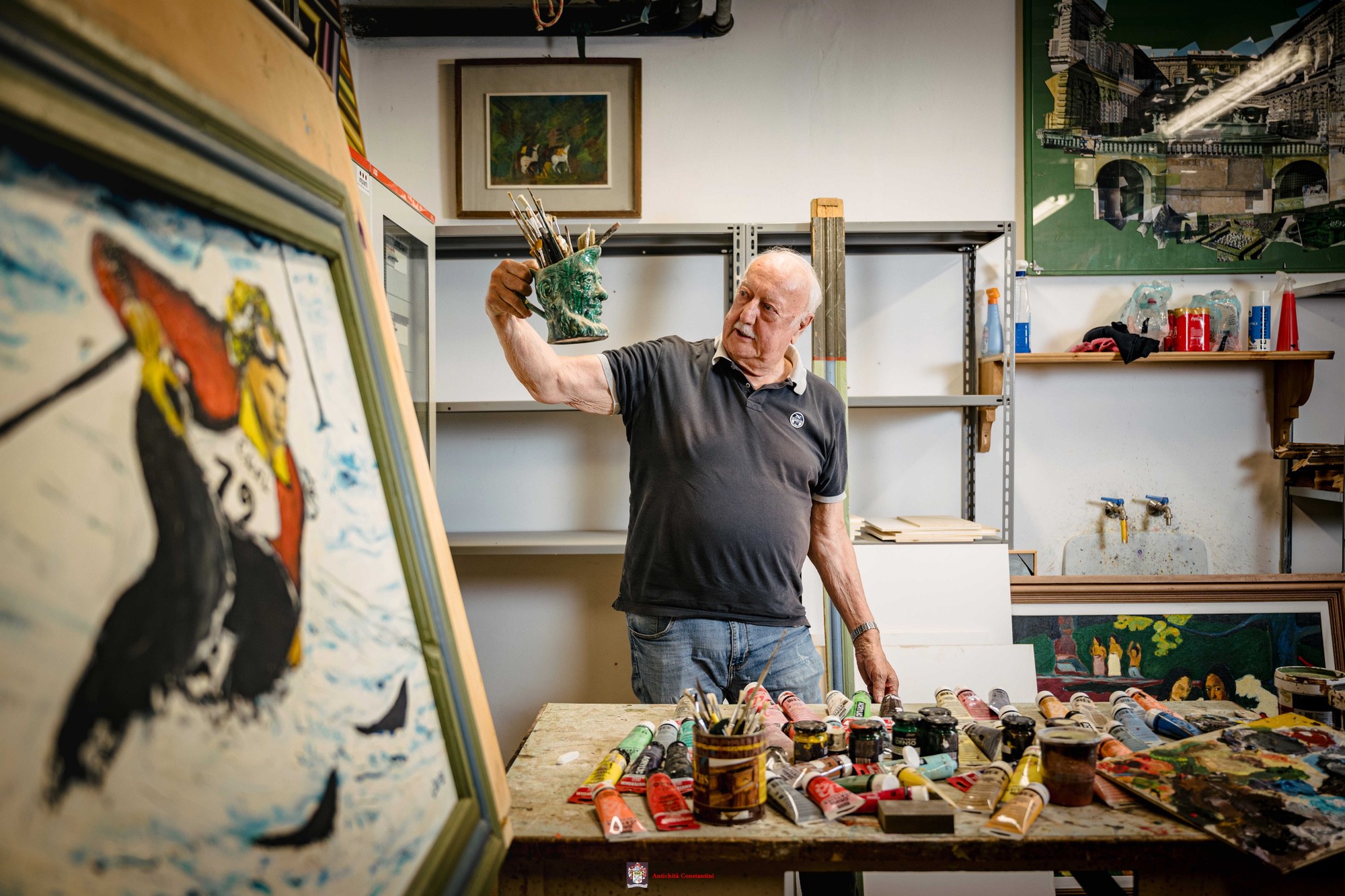 Leopoldo nel laboratorio di pittura Leopoldo solleva un vaso in ceramica verde pieno di pennelli, davanti a un tavolo coperto di tubetti di colore