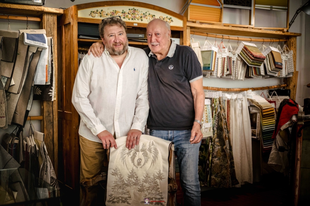 Luca e Leopoldo nella sala tessuti Luca e Leopoldo abbracciati dietro una sedia con schienale ricamato, circondati da campionari tessili appesi