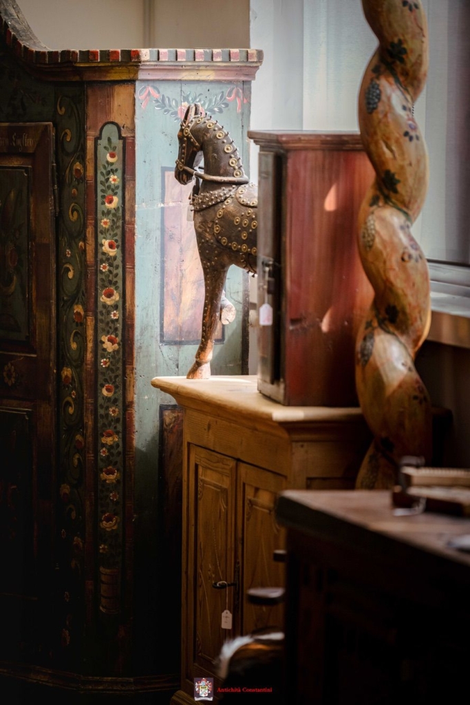 Cavallo in legno tra i mobili dipinti Cavallo scolpito con finiture a borchie su una credenza in legno chiaro, tra un armadio a fasce fiorite e una colonna tortile decorata a grappoli d'uva