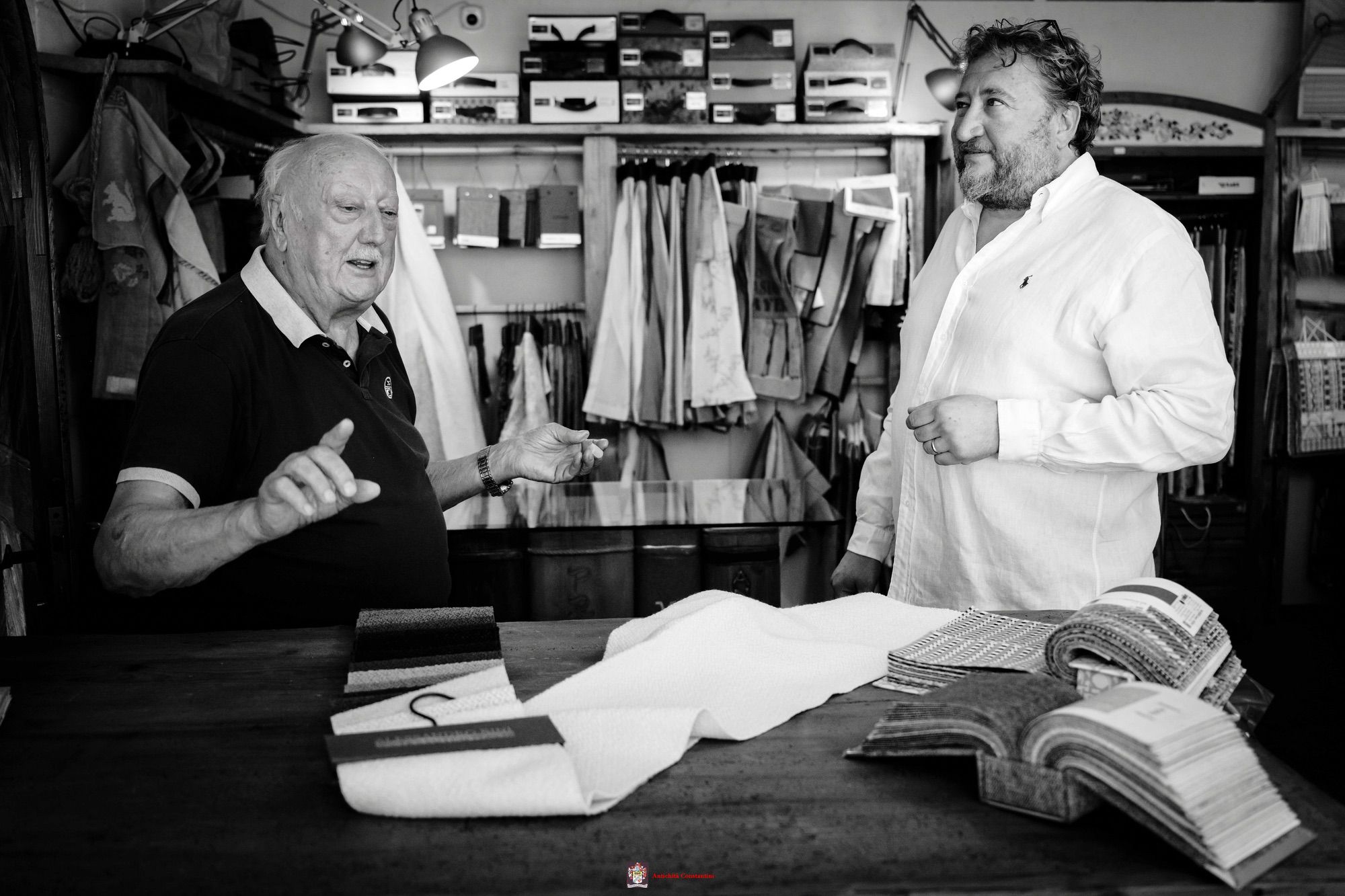 Scatto in bianco e nero: Leopoldo parla gesticolando mentre Luca ascolta, davanti a un bancone con stoffe e cataloghi